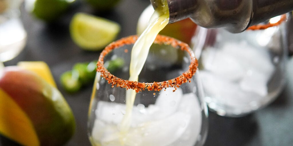 Margarita being poured from shaker into glass with ice and tajin rim