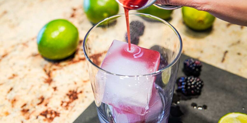 Pouring blackberry simple syrup over ice with limes in the background