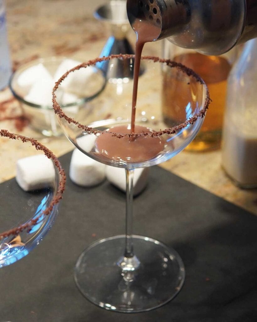 Pouring a smores martini in a chocolate rimmed glass with marshmallows in the background