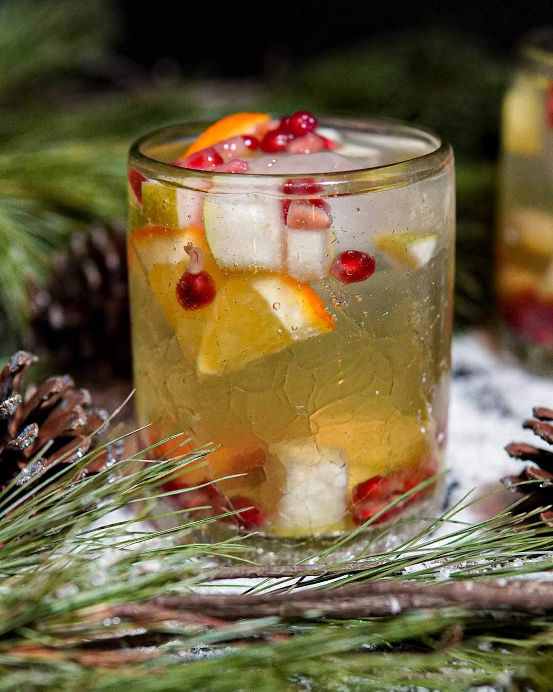 White holiday sangria with orange, pear and pomegranate surrounded by pine, snow and pinecones.