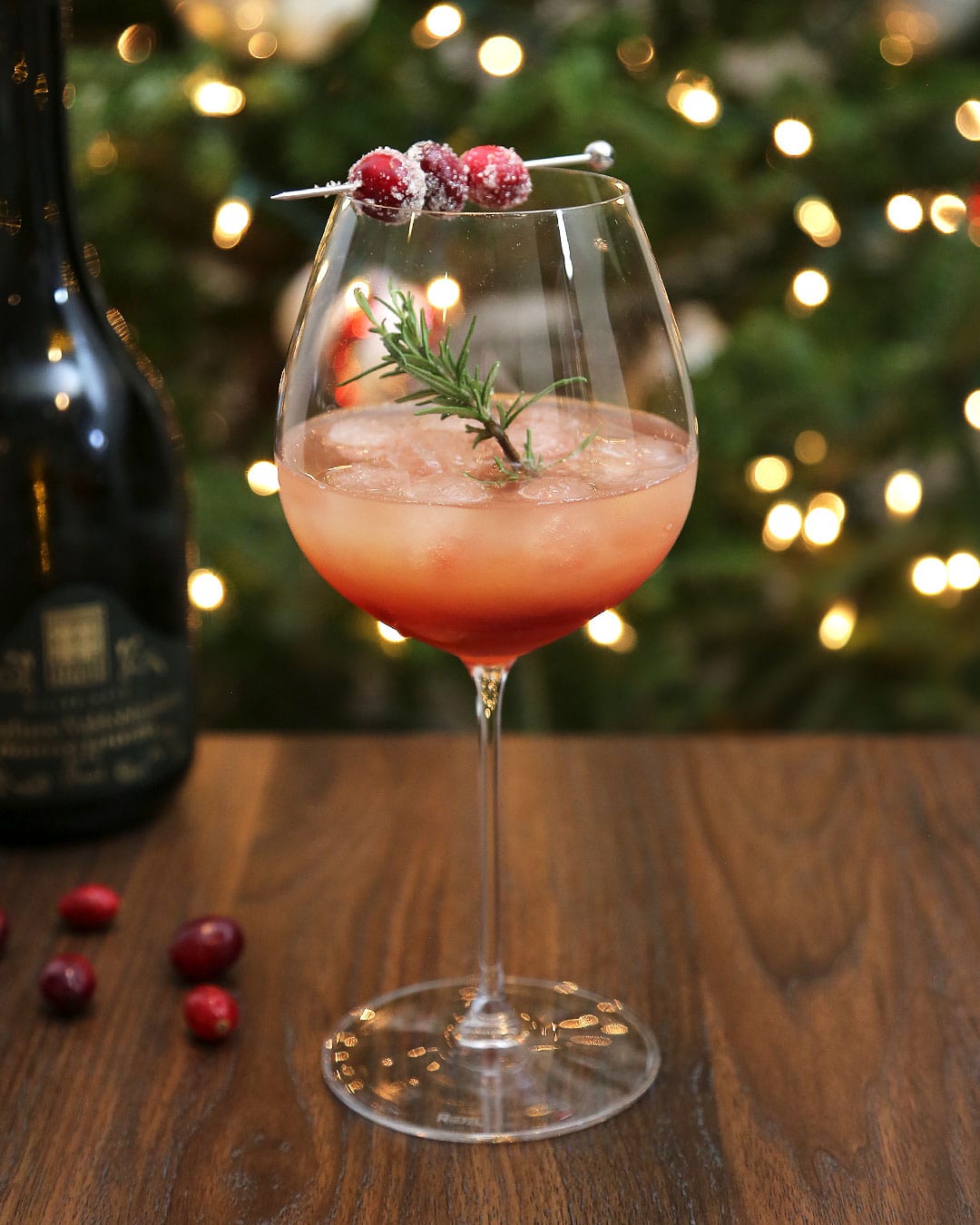 Spiced Cranberry Pear Spritz in wine glass with sugared cranberries and rosemary sprig in front of christmas tree