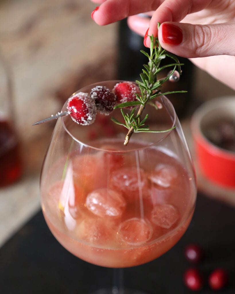 Spiced Cranberry Pear Spritz with sugared cranberries and hand with red nails adding fresh rosemary spring